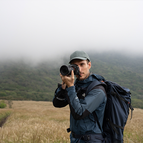 Sony Alpha 7 V Gehäuse - Lifestyle Bild von einem Fotografen der in die Kamera Fotografiert mit Landschaft im Hintegrund