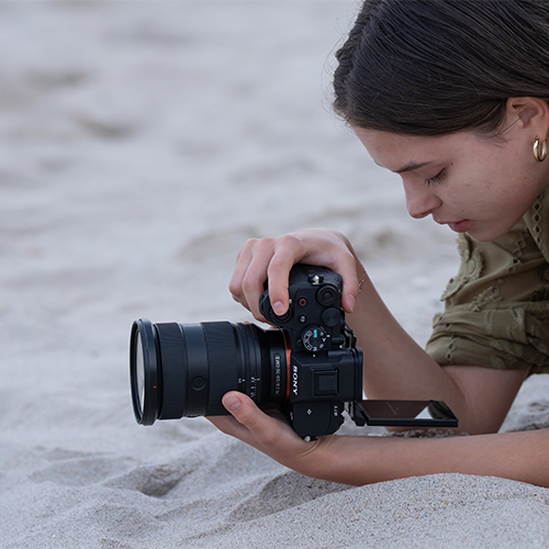 Sony Alpha 7 V Gehäuse - Lifestyle Bild in Verwendung der Kamera, Fotografin liegt am Strand und fotografiert