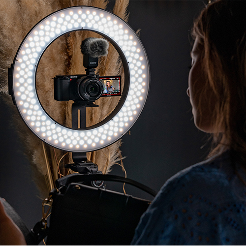 Sony ZV-E10 II Kamera auf einem Stativgriff, von vorne, als Vlogging-Setup mit ausklappbarem Display und externem Mikrofon